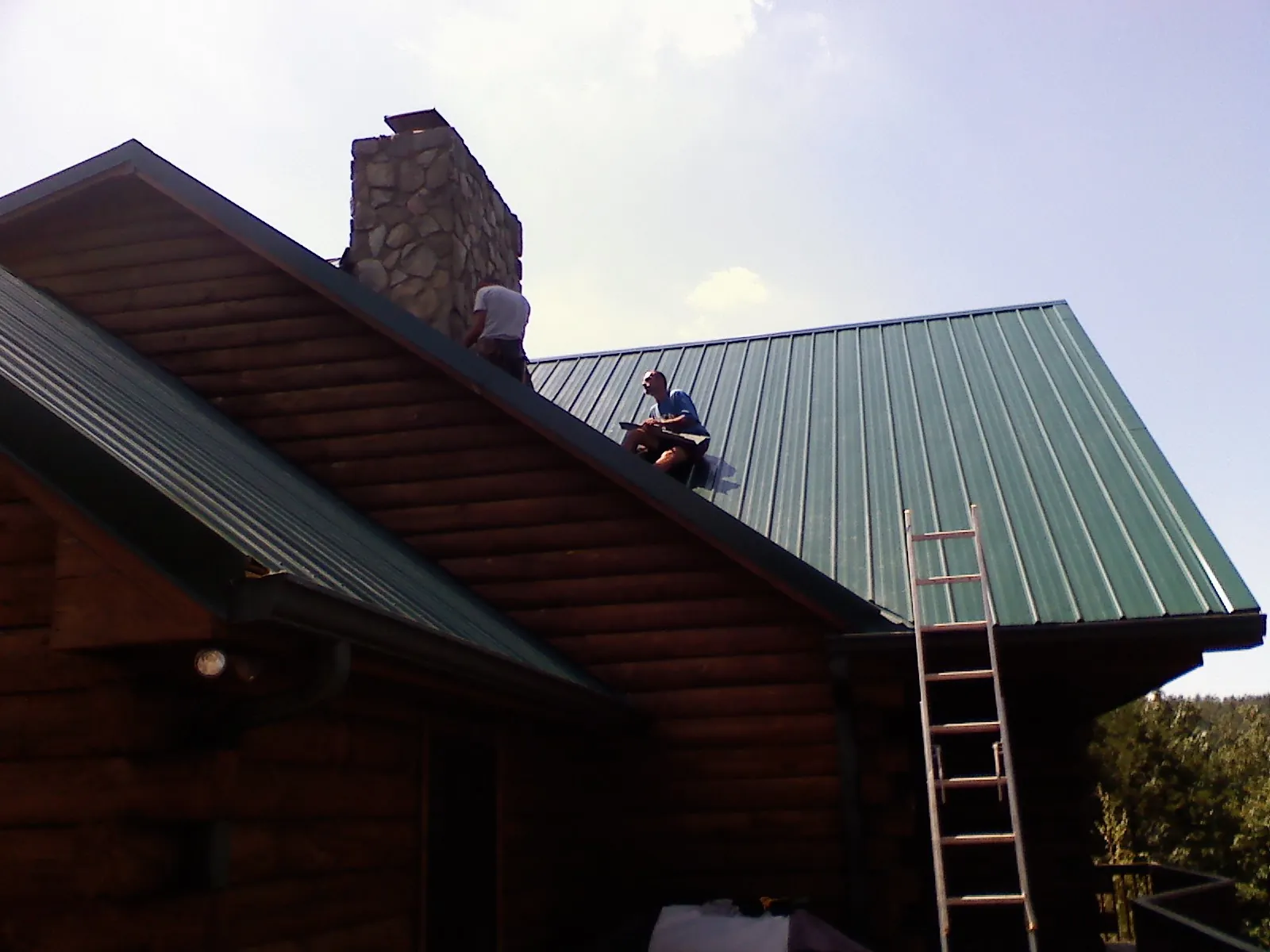 Roofers installing green metal panels beside a stone chimney on a log home in Yorkville